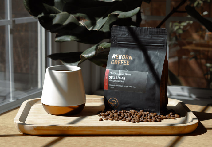 Reborn Coffee bag with coffee beans and a ceramic cup on a wooden tray by a sunlit window.