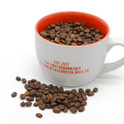 
                  
                    White ceramic cup with orange interior, has text that reads, “Est, 2017, the first reborn cafe, village of la floresta, brea, C,A.” Has a handle. Filled with coffee beans. 3 of 4.
                  
                