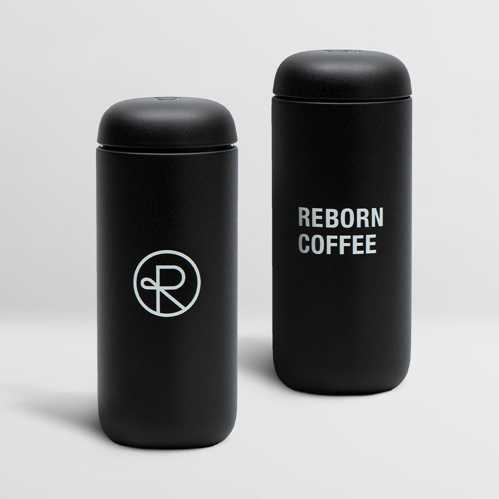 Two black colored mugs. One mug has the Reborn logo, the other mug has the words Reborn Coffee stacked vertically. Leads to online store page for Reborn Coffee mugs and kettles.