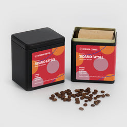 
                  
                     Two black metal boxes, one opened with Ethiopia Sidamo Faysel Coffee beans inside. Sticker shows product info imposed on circular design. Front view, 4 of 4.
                  
                
