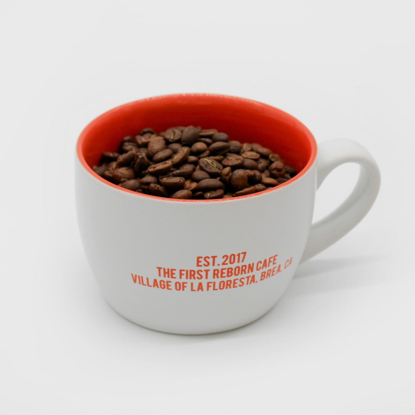 White ceramic cup with orange interior, has text that reads, “Est, 2017, the first reborn cafe, village of la floresta, brea, C,A.” Has a handle. Filled with coffee beans. 2 of 4.