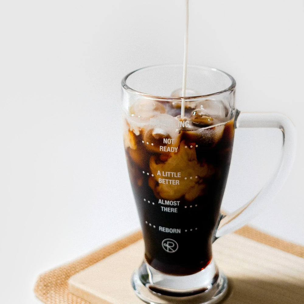 Reborn Coffee pint glass with text that reads from top to bottom, “I’m feeling, Not ready read, a little better, almost there, Reborn.” Glass is filled with coffee and milk is being poured in. 1 of 4.