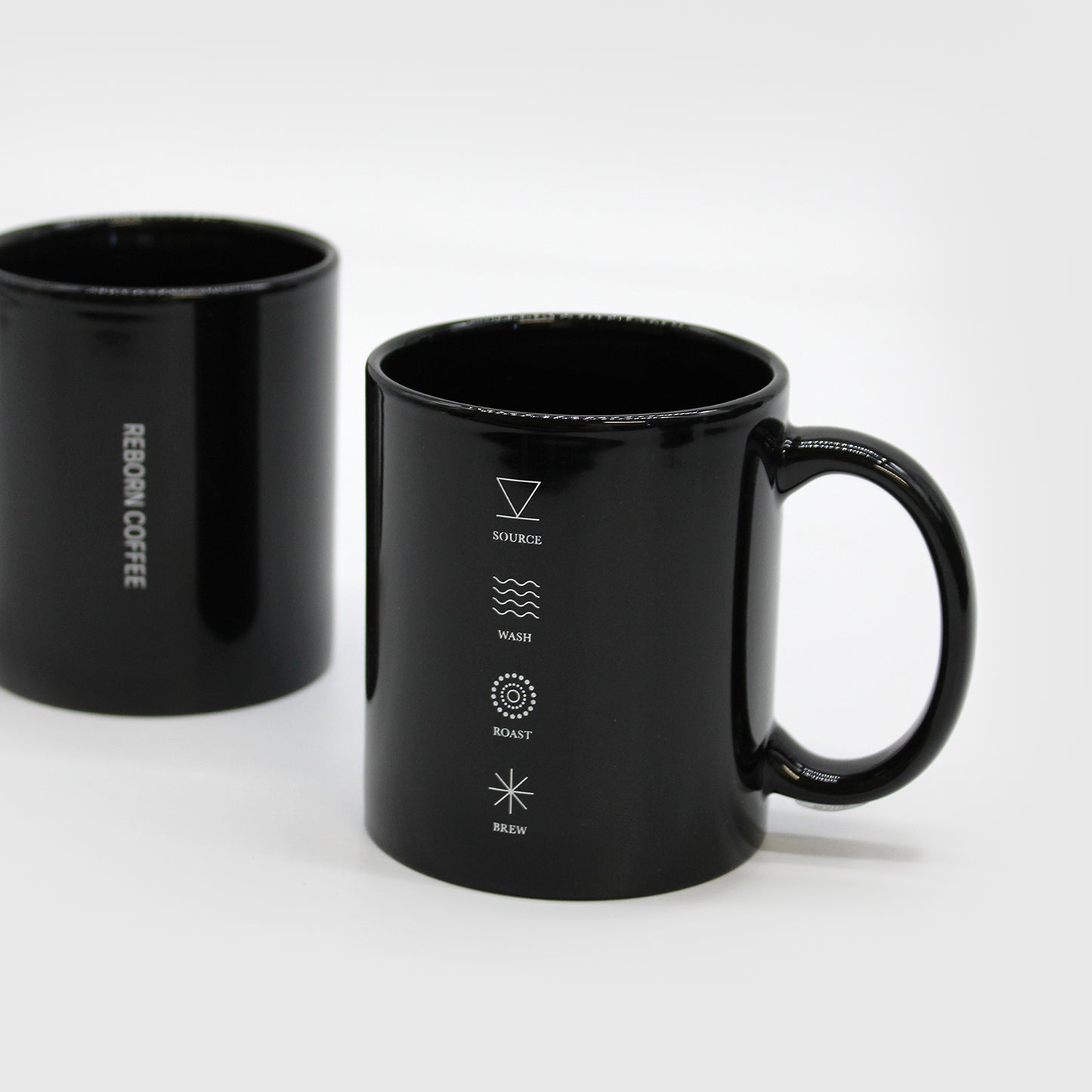 Black Ceramic mug with C handle, has 4 symbols aligned vertically, with text under each symbol that reads, “Source, Wash, Roast, Brew.” Second mug to the left has text, “Reborn Coffee,” on it. 2 of 3.