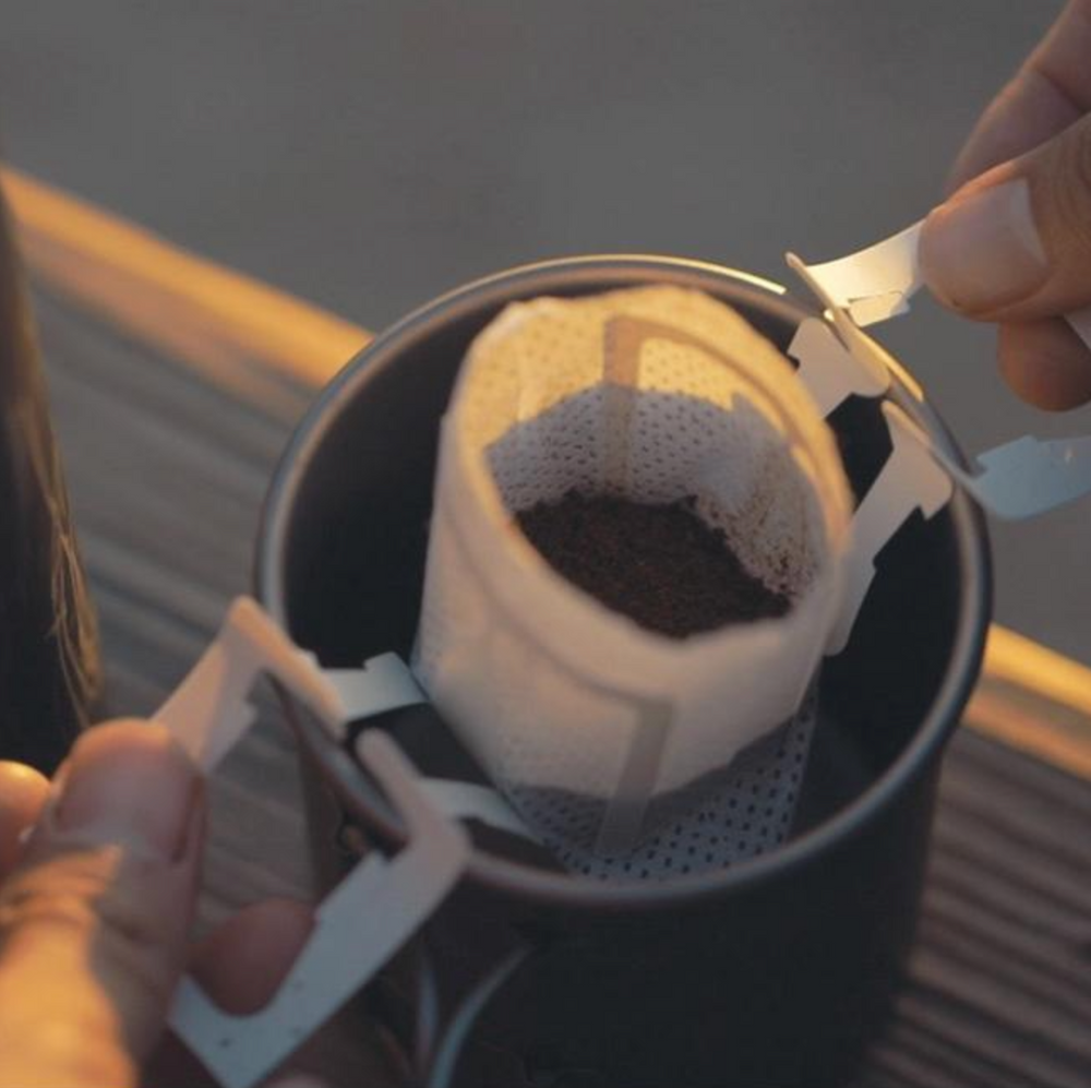 
                  
                    Pour over pack, hanging over mug, with hanging paper arms. Grounded coffee is shown. 
                  
                